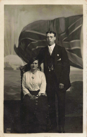 Old real photo postcard of a lady and gentleman standing in front of a British flag